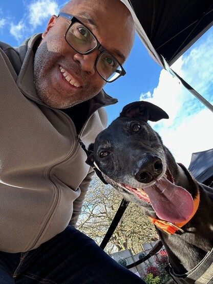 A view from below of a smiling human taking a selfie with a black greyhound boy. The hound's jaws are open and his tongue is lolling out.