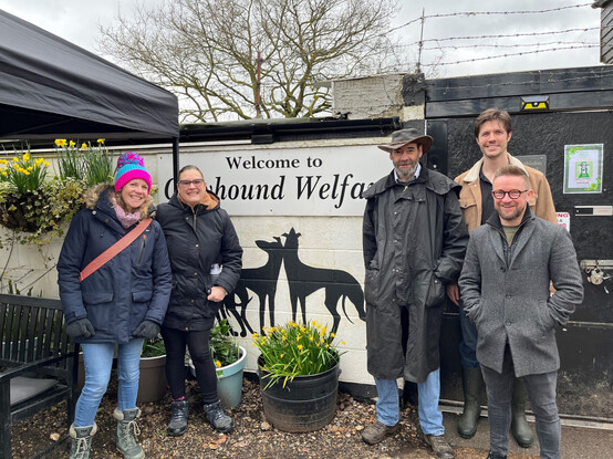 A photos of a group of 5 humans standing outside in the kennel yard. Behind them is a white wall with a silhouette of two greyhounds and the words "Welcome to Greyhound Welfare"