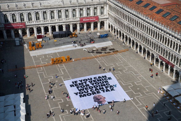 An aerial photo of a huge banner unfurled in Venice's Piazza San Marco. The text on the banner is exactly as the post describes, in black capital letters with an image of Jeff Bezos's laughing face below.