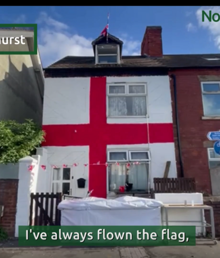 A screenshot of a news report showing a frontal view of a left end terrace two storey house with small front garden, white wall and black wooden gate, The house is painted white with the red and white cross of St George across the en tirety of the front of the house from gutter line to ground. A caption at the bottom  reads, "I've always flown the flag,"
